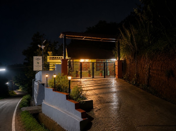A gated entrance to a property at night with a paved driveway leading up to a covered area with steps and potted plants.