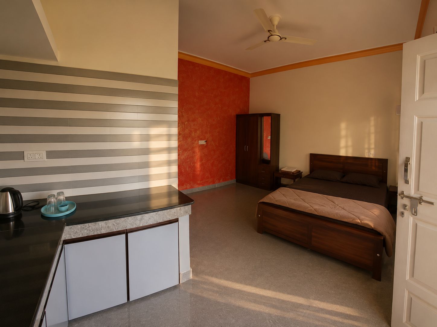 A bedroom with a bed, a wardrobe, a desk with a kettle, and a ceiling fan.