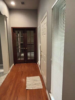 A hallway with a wooden floor, a white rug, a door with glass panels, and a window with blinds.