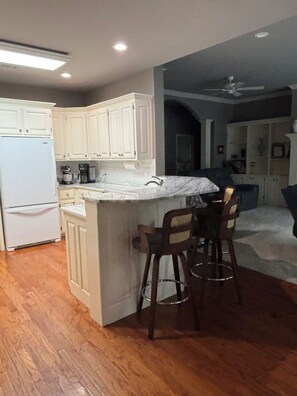 A kitchen with white cabinets, a white refrigerator, a marble countertop, and a bar area with stools.