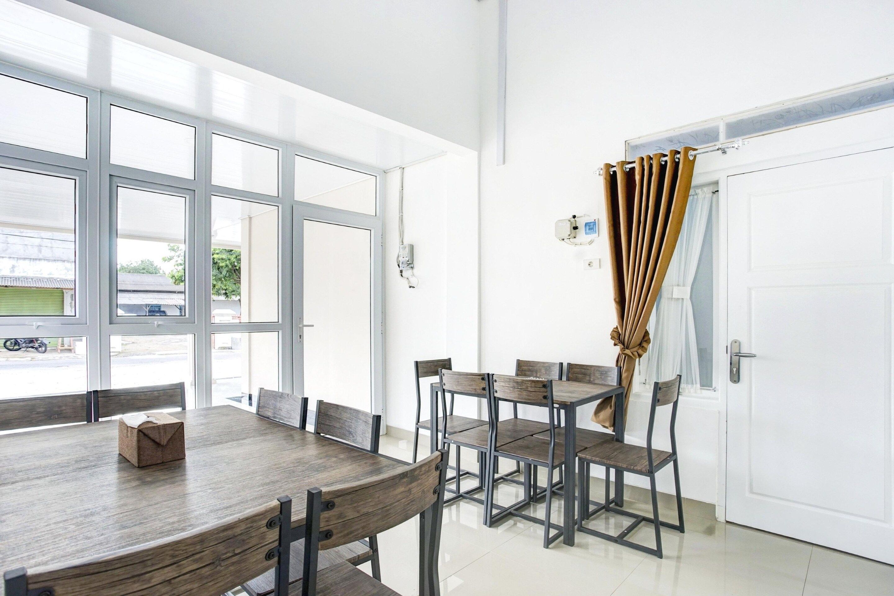 A dining area with a large table and chairs, a white door with a curtain, and a view through the windows.