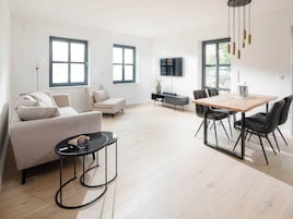 Una sala moderna con un sofá, una mesa de comedor y sillas. Hay un televisor montado en la pared y un ventanal que deja entrar luz natural.