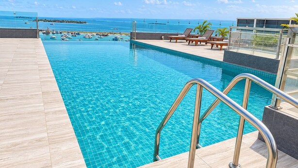 Piscina con vista al mar y barcos, rodeada de sillas de descanso y un edificio moderno.