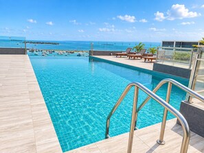 A swimming pool with a view of the sea and boats, surrounded by lounge chairs and a modern building.