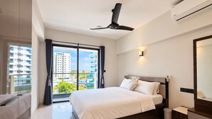 Habitación de hotel moderna con una cama grande, un ventilador de techo, un equipo de aire acondicionado, un baño con inodoro y lavamanos, y un ventanal con vistas a edificios y zonas verdes.