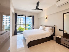 Habitación de hotel moderna con una cama grande, un ventilador de techo, un equipo de aire acondicionado, un baño con inodoro y lavamanos, y un ventanal con vistas a edificios y zonas verdes.