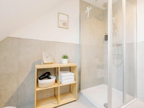 Ein modernes Badezimmer mit einer Duschkabine aus Glas, einem Holzregal mit Handtüchern und einem Fön, sowie einer kleinen Topfpflanze.