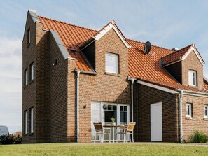 Ein zweistöckiges Backsteinhhaus mit Ziegeldach, einer Haustür und einer kleinen Terrasse mit Tisch und Stühlen.