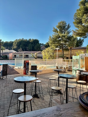An outdoor seating area with round tables and stools, a pool in the background, and a building with a tiled roof. - Studio for 4 people – Sanary-sur-Mer – 10 minutes’ walk from the beach (Sanary-sur-Mer)
