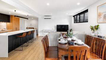 A modern kitchen with a central island, a dining area with wooden chairs, and a flat-screen TV mounted on the wall.