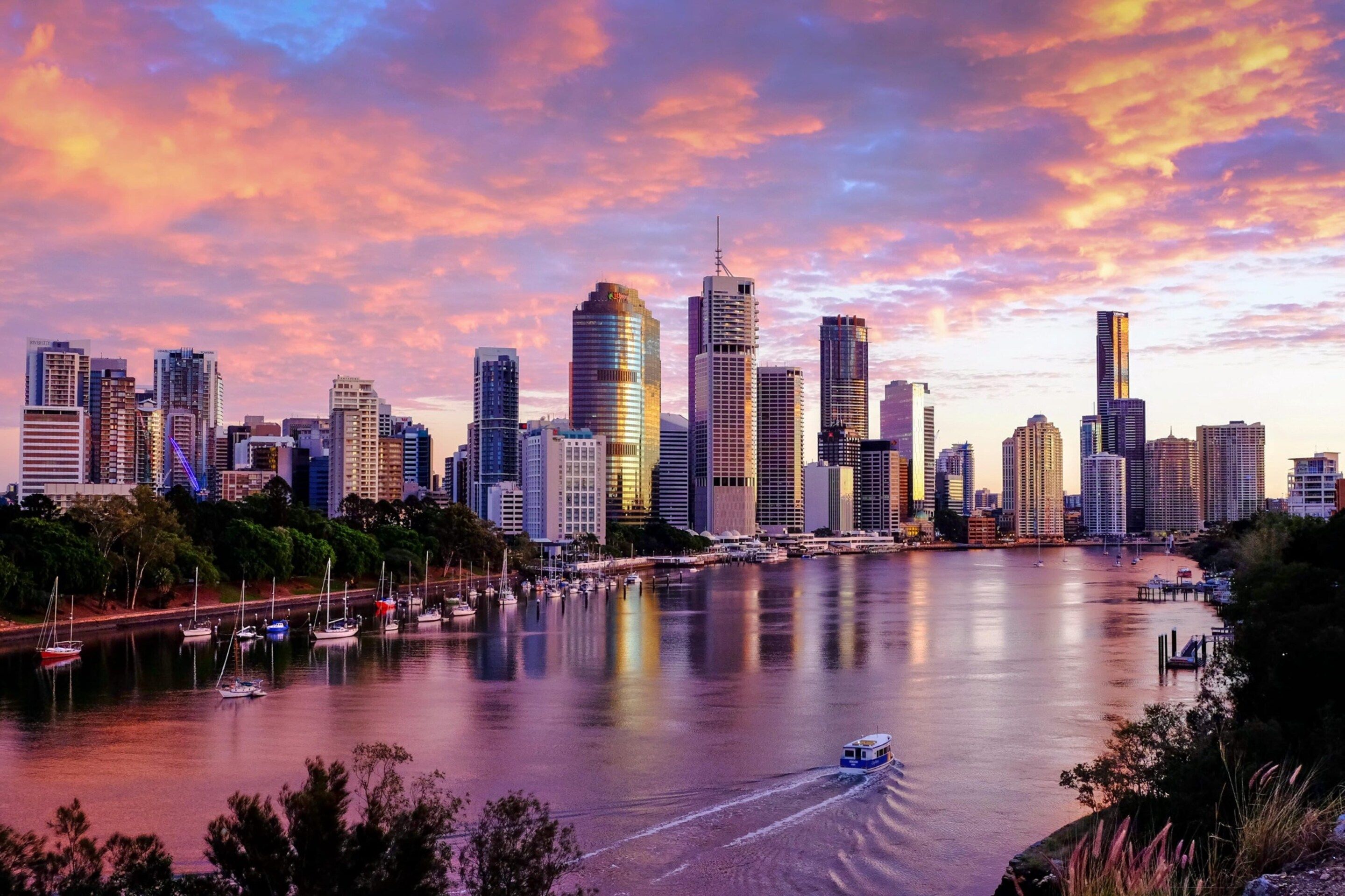 A city skyline with high-rise buildings, a marina with boats, and a river reflecting the sunset.