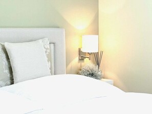 Ein ordentlich bezogenes Bett mit weißen Leinen und einem Kopfteil, ein Nachttisch mit Lampe und Schilferdiffusor sowie eine Wandleuchte.