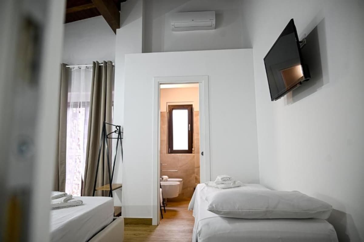 Habitación de hotel con cama, televisor montado en la pared, baño visible a través de una puerta abierta y ventana con cortinas.