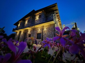 A two-story stone building with balconies, illuminated windows, and a garden with purple flowers in the foreground. - Borgo Caridà - Green Family Holidays (Zaccanopoli)