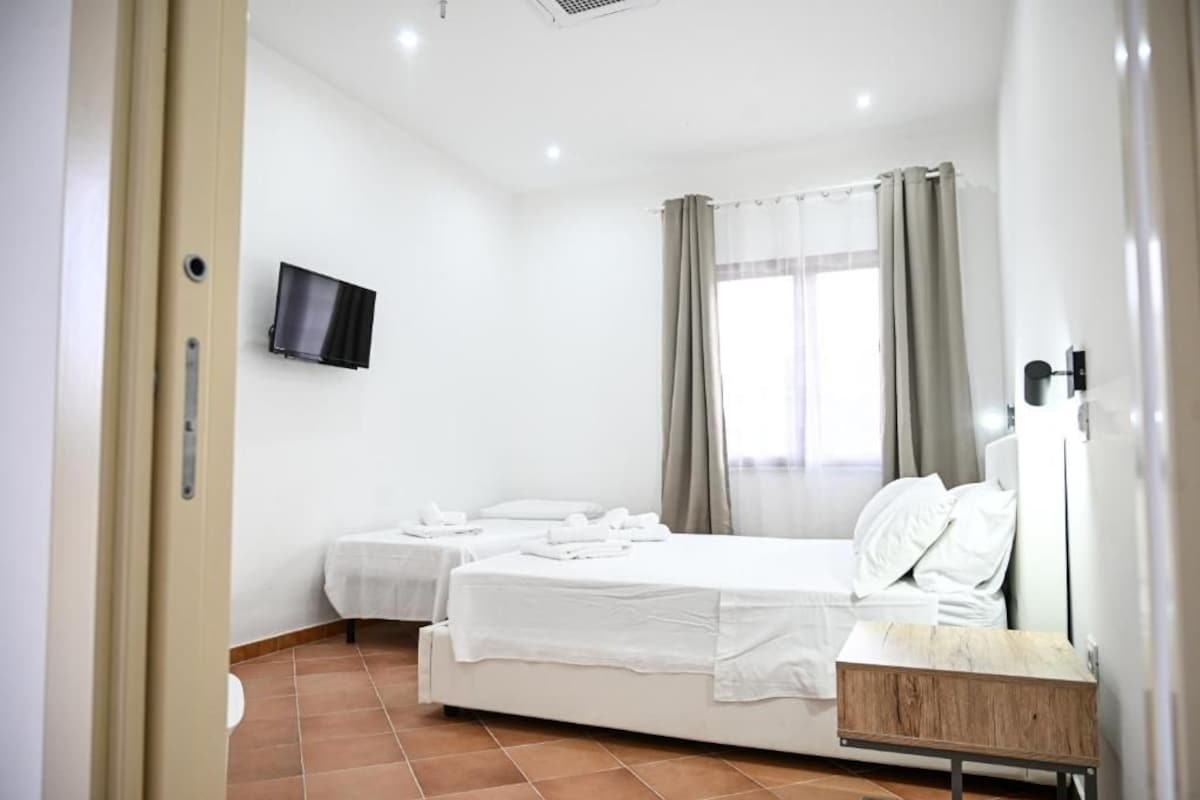 Una habitación de hotel con dos camas, un televisor instalado en la pared, una ventana con cortinas y una pequeña mesa de madera.