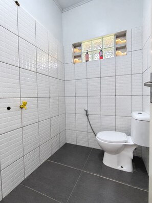 Baño con paredes de azulejos blancos, inodoro blanco, zona de ducha y una pequeña ventana con detalles decorativos.