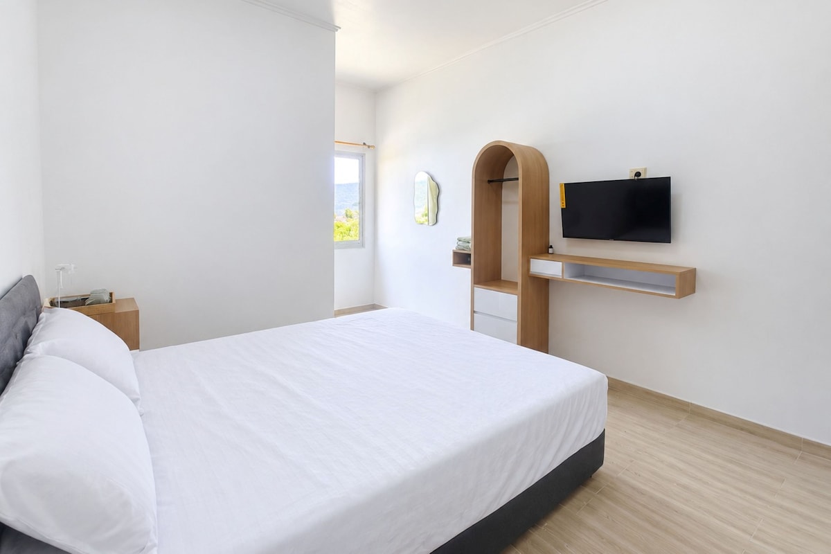 Moderni hotellihuone, jossa on suuri sänky, seinälle asennettu televisio ja ikkuna, josta on näkymä ulos.
