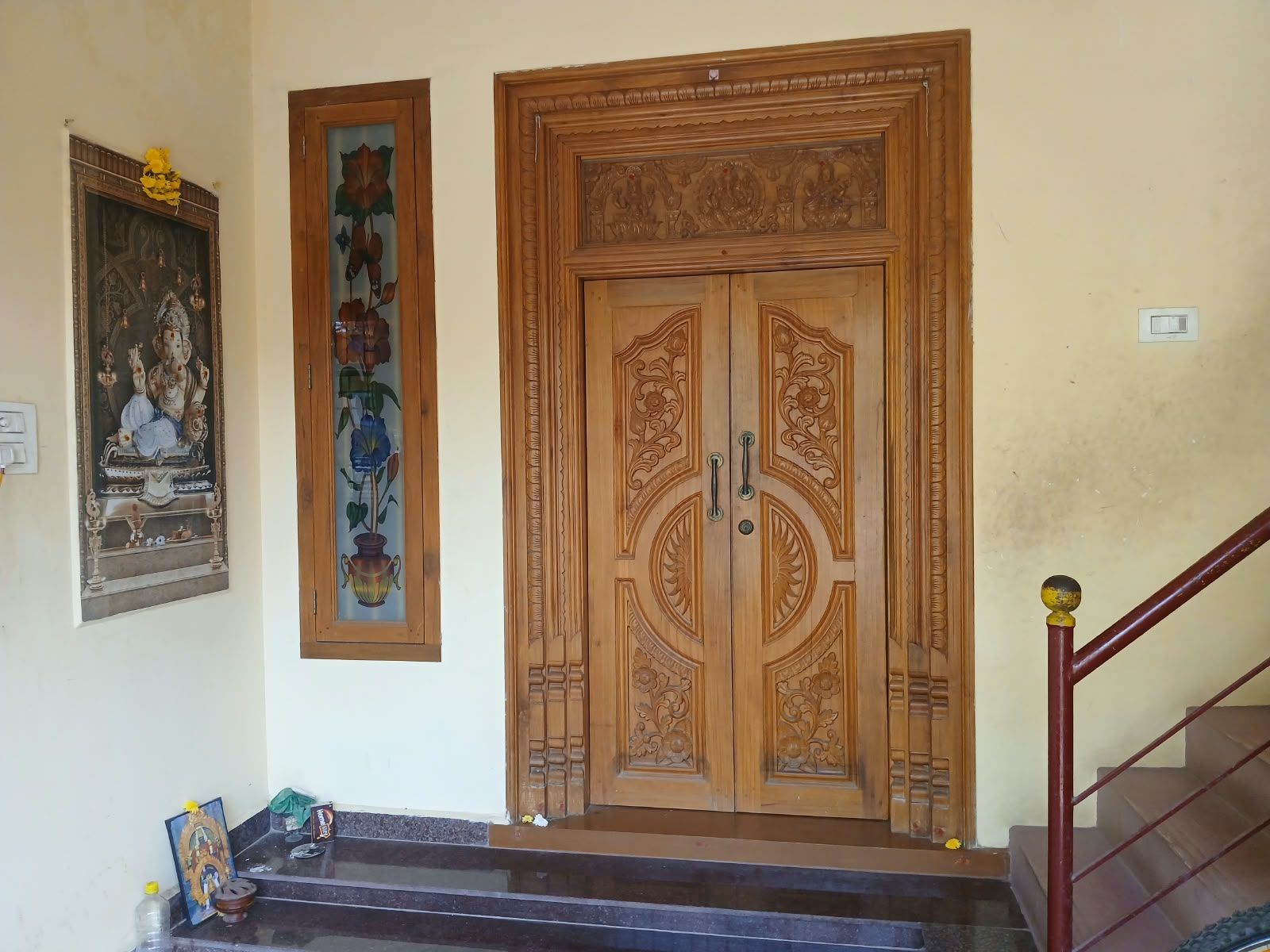 A wooden double door with intricate carvings, a framed painting of a deity on the wall, and a staircase with a handrail.