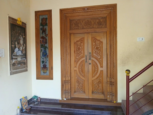 A wooden double door with intricate carvings, a framed painting of a deity on the wall, and a staircase with a handrail.