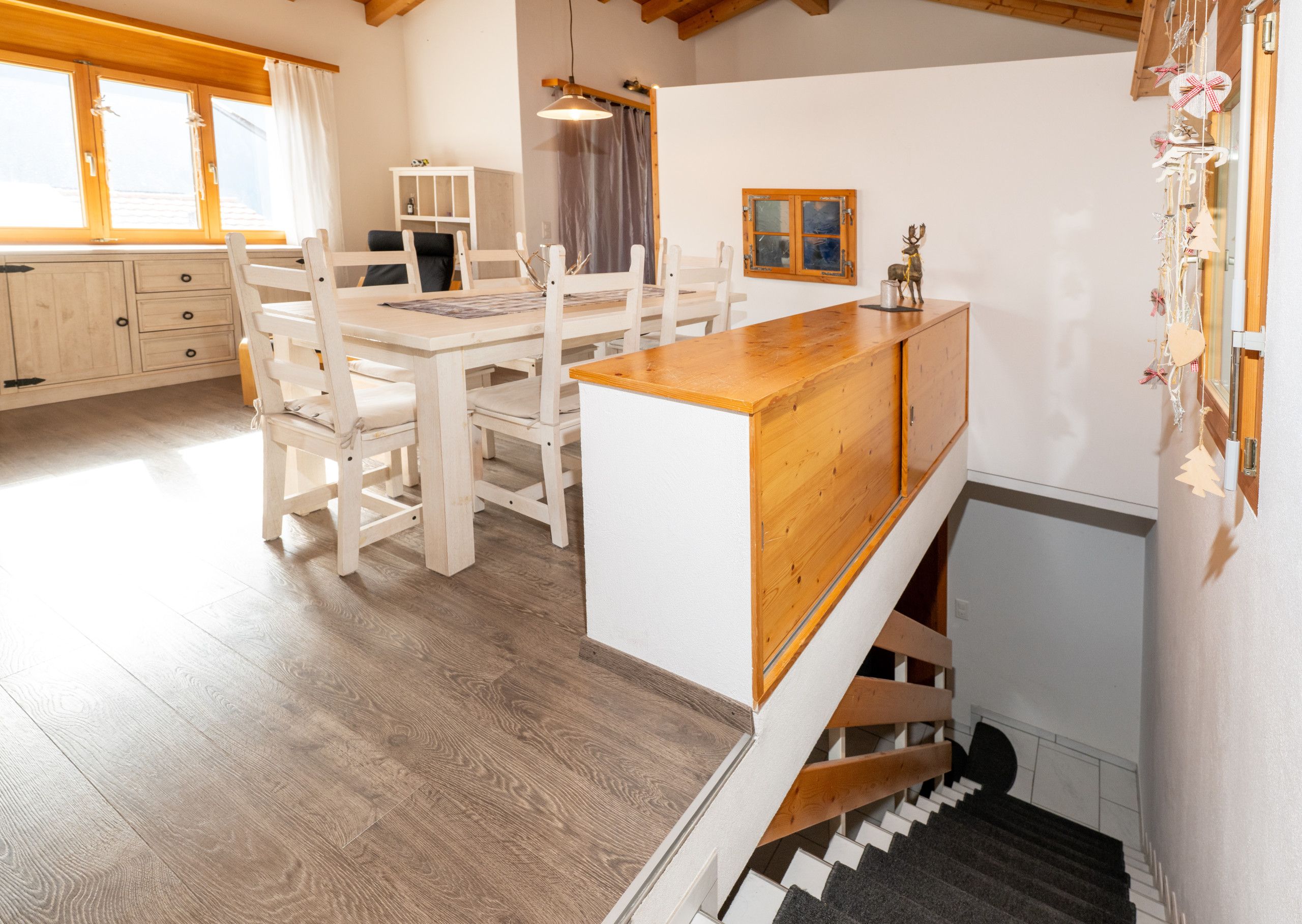 A wooden dining table with chairs, a staircase with a wooden railing, and a white wall with a picture frame.
