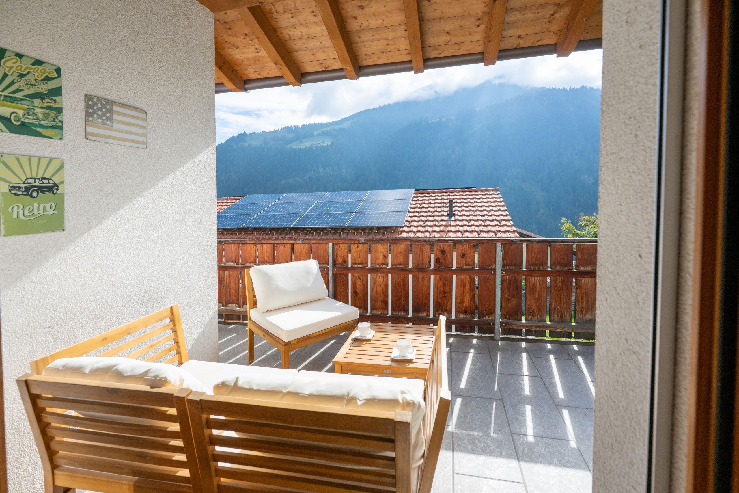 Een balkon met een houten stoel en tafel, uitzicht op bergen, en zonnepanelen op een gebouw.
