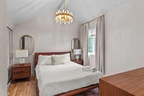 A bedroom with a large bed, two bedside tables with lamps, a wooden headboard, and a chandelier.
