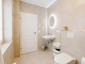 A compact bathroom with a white door, a sink, a toilet, and a small trash bin. - Holiday Apartment in Greetsiel With Wi-fi (Krummhoern)