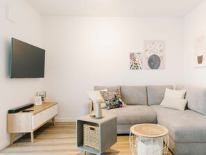 Een moderne woonkamer met een grijze bank, een klein bijzettafeltje, een flatscreen televisie aan de muur en een houten vloer.