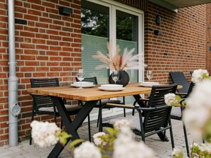 A brick exterior wall with a window and a door, a wooden table with chairs, and a vase with dried pampas grass. - Holiday Apartment in Greetsiel With Wi-fi (Krummhoern)