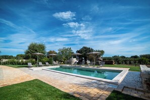 A rectangular swimming pool with a tiled deck, surrounded by lounge chairs and umbrellas, set against a backdrop of trees and a clear sky. - Trullo Sippuddo (Puglia)