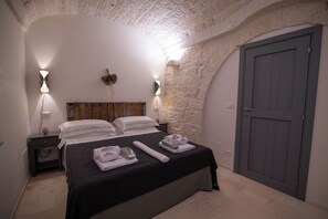 A bedroom with a stone wall, a wooden headboard, a bed with white linens, two bedside lamps, and a door. - Trullo Sippuddo (Puglia)