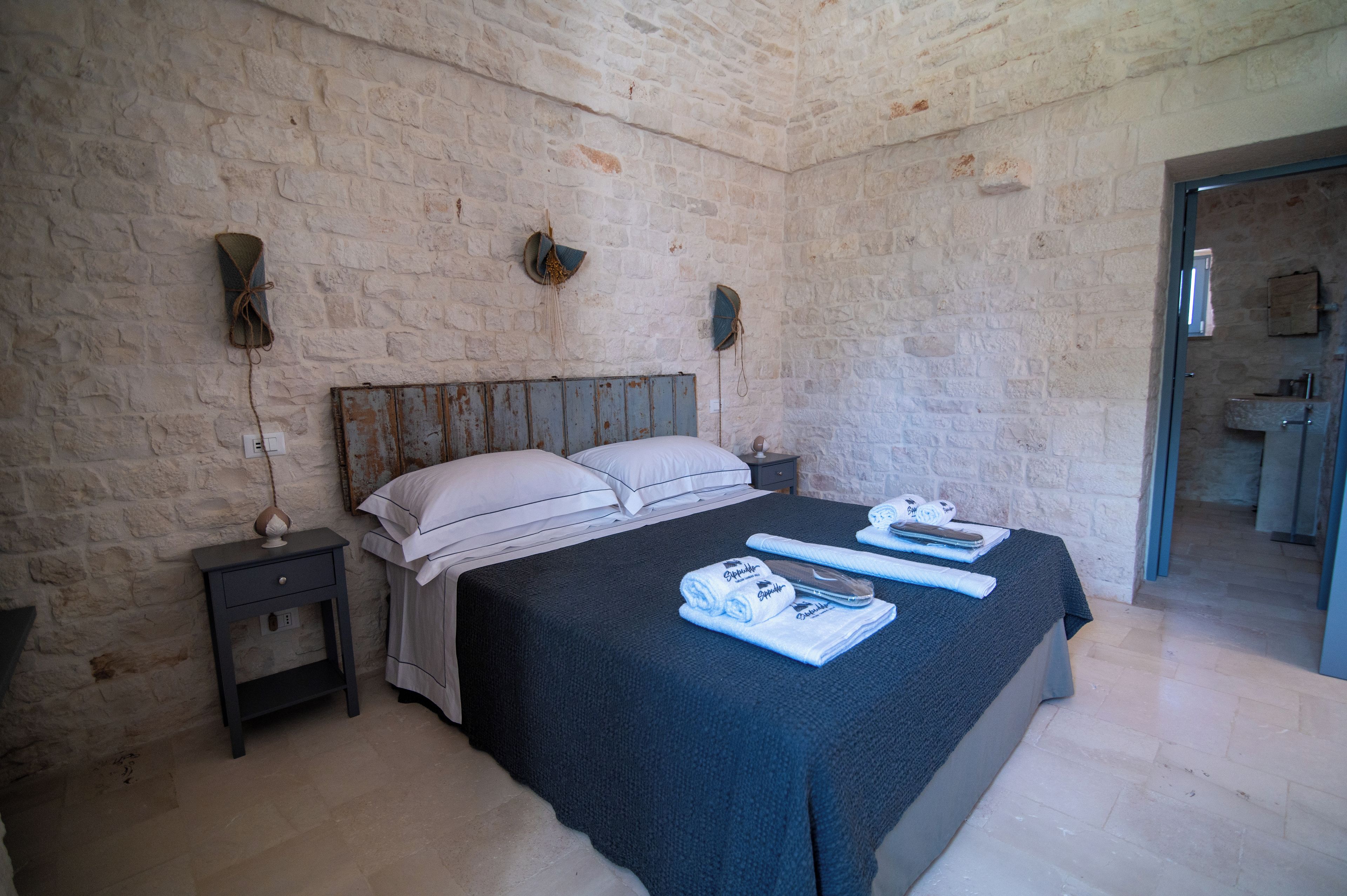 A bedroom with a stone wall, a wooden headboard, a bed with white linens, a nightstand with a lamp, and a bathroom visible through an open door.