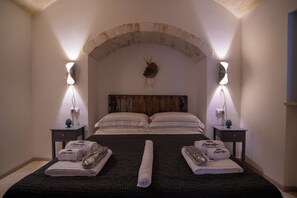 A bedroom with a stone arch ceiling, a wooden headboard, two bedside tables with lamps, and a bed with rolled towels and a rolled-up blanket. - Trullo Sippuddo (Puglia)