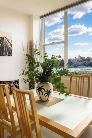 Ein Essbereich mit Tisch, Stühlen, einer Pflanzvase, einem Bild an der Wand und Blick auf eine Stadtlandschaft durch ein großes Fenster.