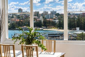 Ein Zimmer mit einem großen Fenster mit Blick auf eine Stadtlandschaft, einem Essbereich mit Stühlen und einem Tisch und einer Zimmerpflanze im Vordergrund.