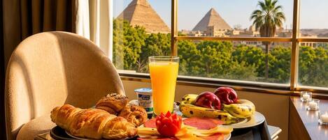 Un vassoio colazione con croissant, insalata di frutta, un bicchiere di succo d'arancia e un piatto di prosciutto e formaggio, con vista sulle piramidi sullo sfondo.