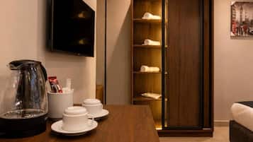 A hotel room with a wooden dining table, a coffee maker, a flat-screen TV, a wardrobe with shelves, and a painting on the wall.