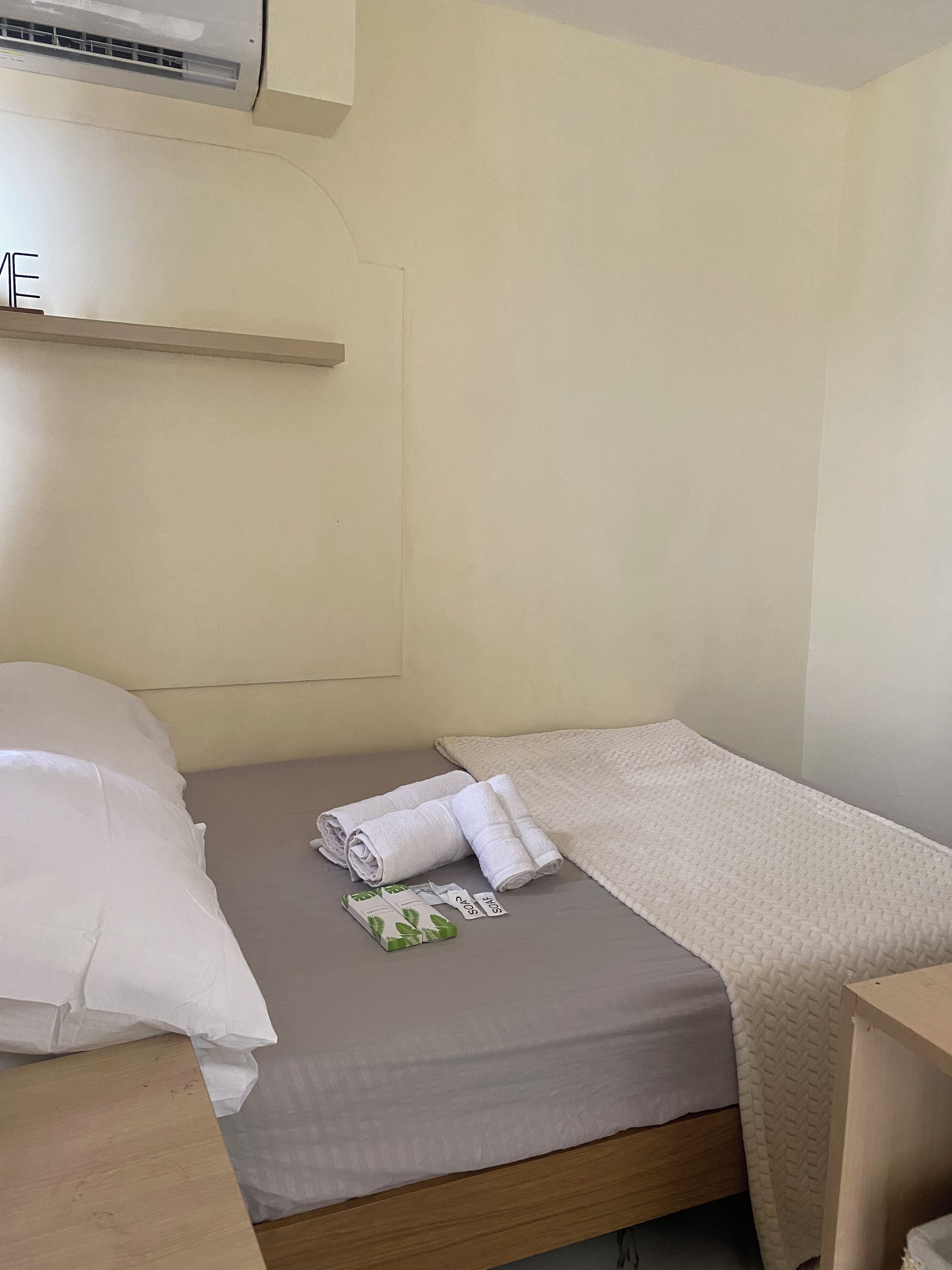 A single bed with a beige blanket and rolled towels.