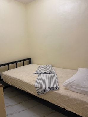 Uma cama de solteiro com colchão branco e manta listrada, travesseiro branco e cabeceira preta em quarto com paredes lisas e piso de azulejos.