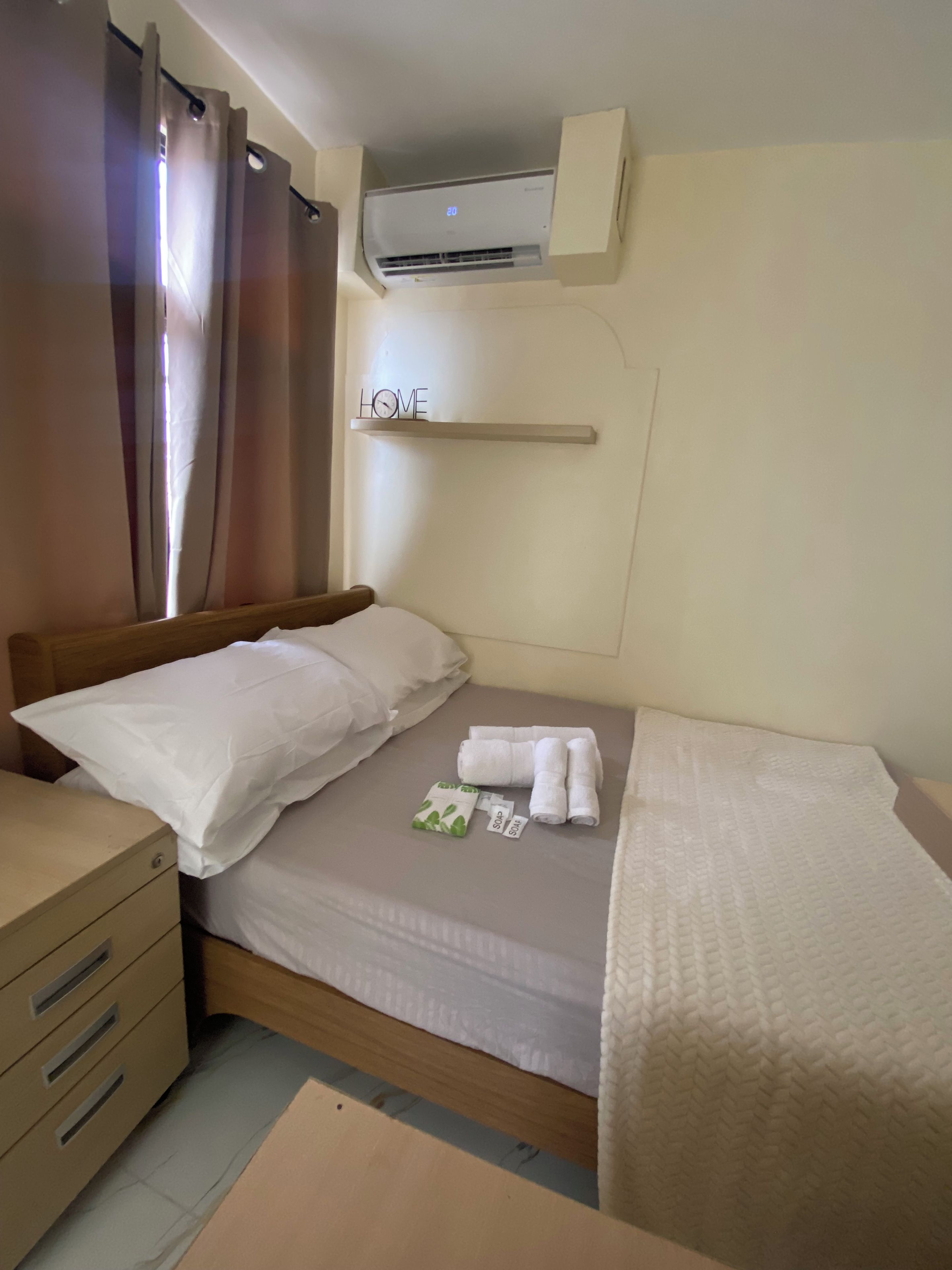 Hotelkamer met een bed, nachtkastje, ladeblok, airconditioning en een plank met een 'HOME' bordje.