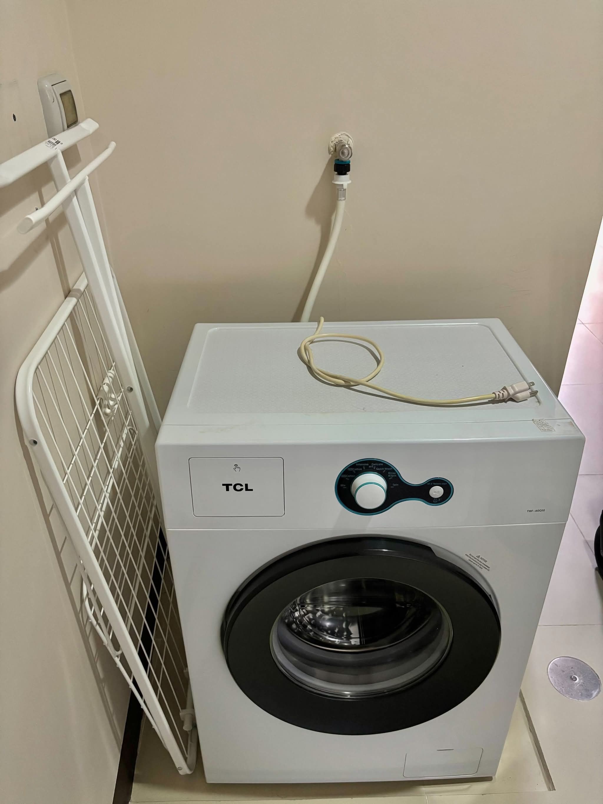 A white TCL washing machine with a front-loading door, a control panel with a dial, and a power cord plugged in.