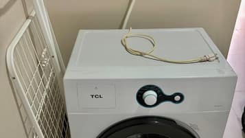 A white TCL washing machine with a front-loading door, a control panel with a dial, and a power cord plugged in.