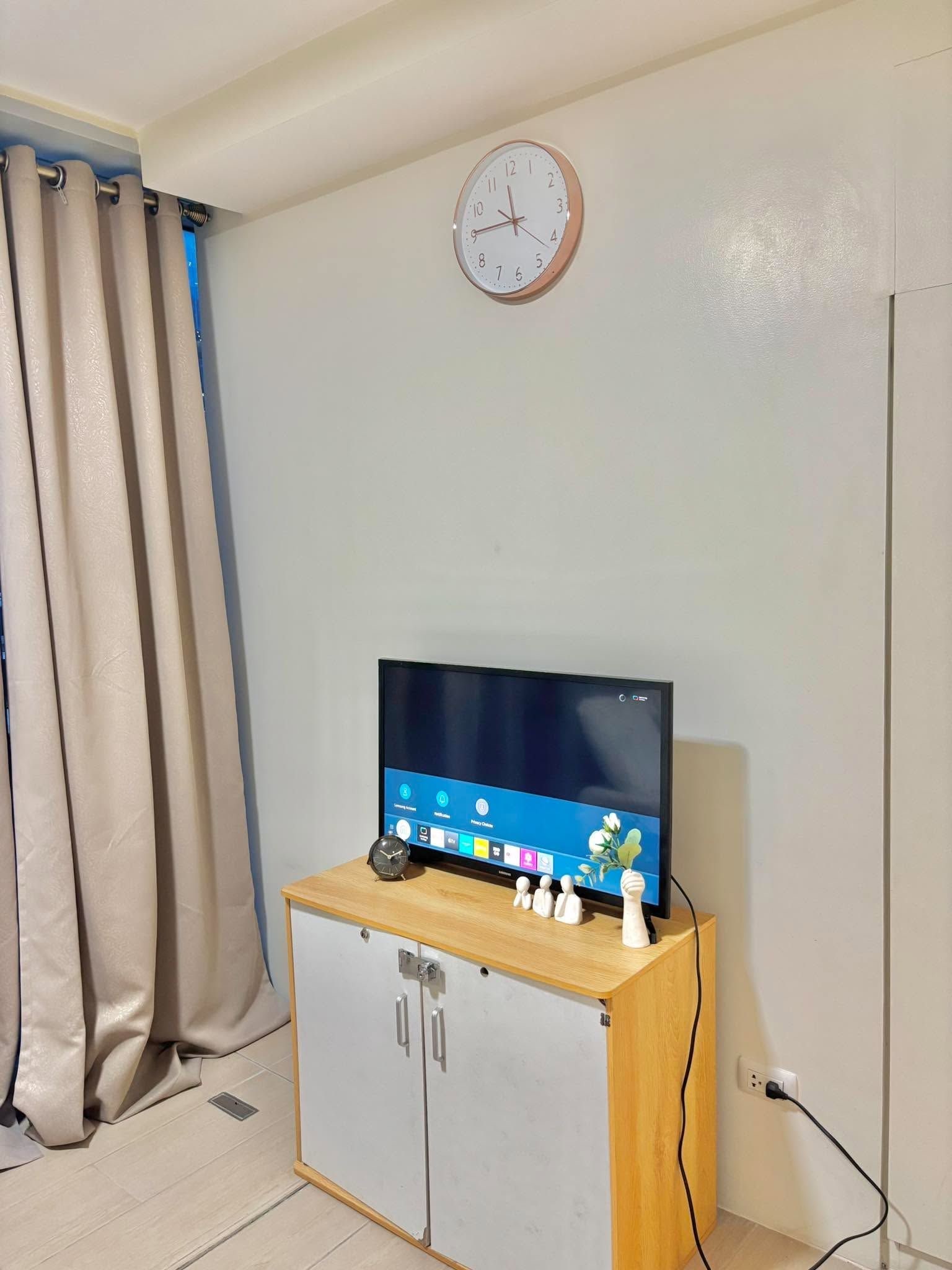 A room with a television on a wooden cabinet, a clock on the wall, and a curtain on the window.