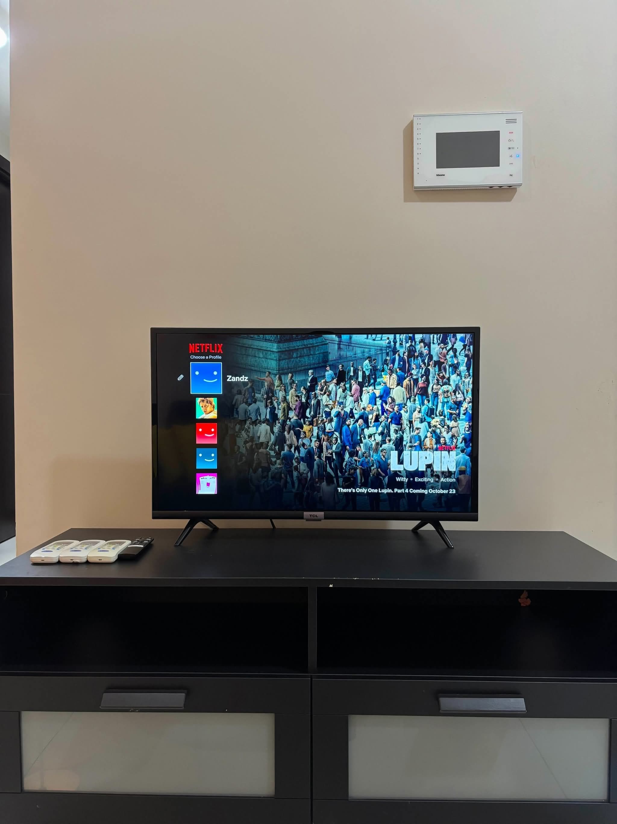 Un televisor sobre un soporte negro, con la interfaz de Netflix en la pantalla, un termostato blanco arriba y un mueble de entretenimiento negro debajo.