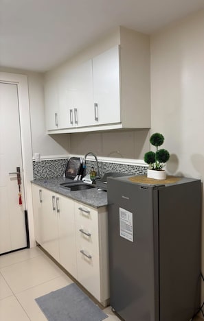 A compact kitchen with white cabinets, a granite countertop, a built-in sink, and a small refrigerator. - COZI Uptown Parksuites (Taguig)