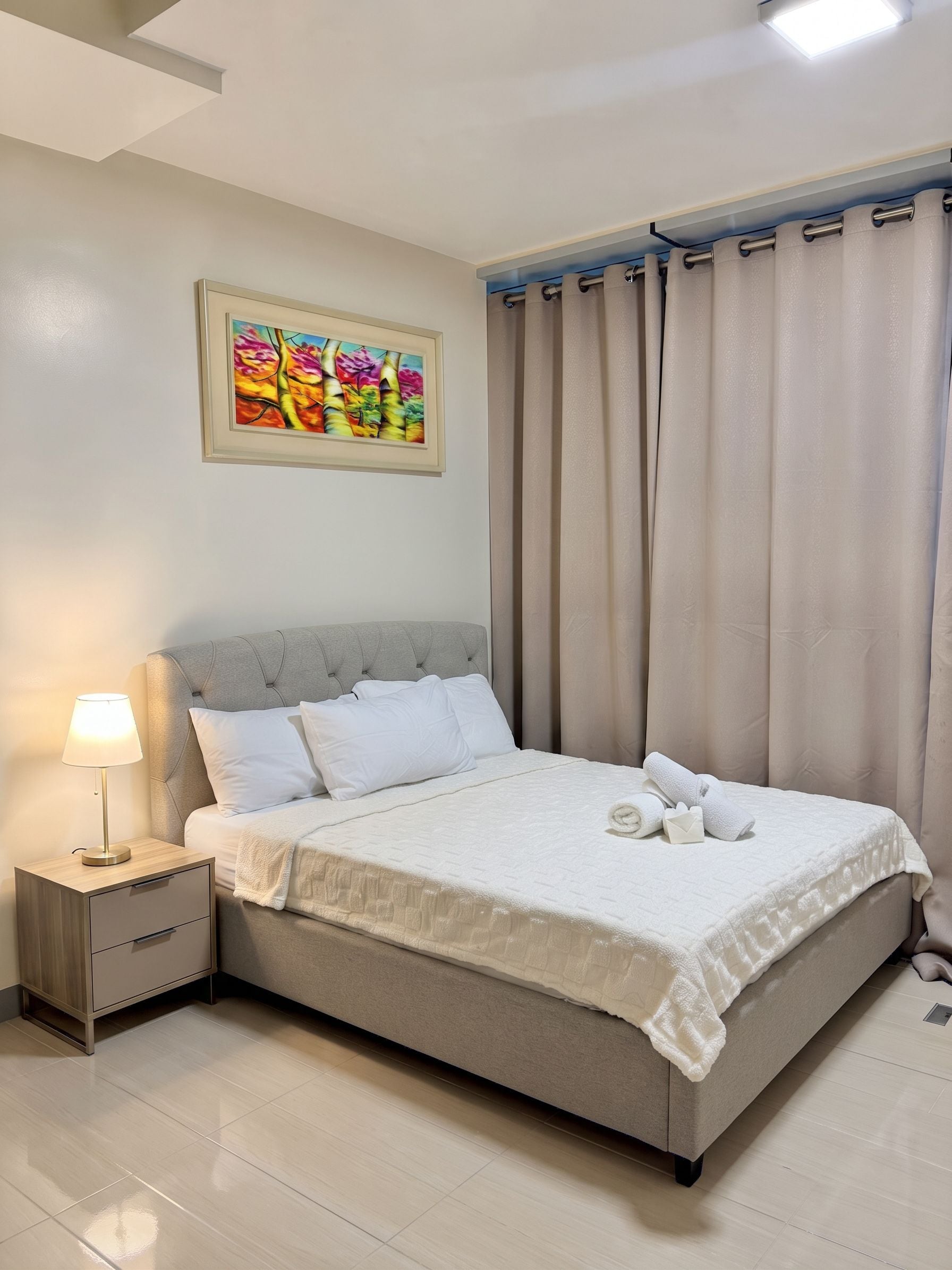 Una cama bien hecha con sábanas blancas, una lámpara de noche y un cuadro en la pared.