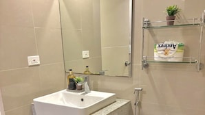 Un baño con lavamanos blanco, espejo, inodoro y ducha. Hay un estante pequeño con una planta y una caja de pañuelos.