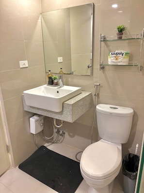 A bathroom with a white sink, mirror, toilet, and shower. There is a small shelf with a plant and a box of tissues. - COZI Uptown Parksuites (Taguig)