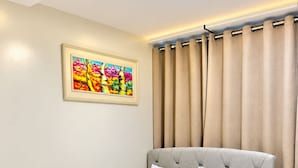 Una cama bien hecha con ropa de cama y almohadas blancas, un cuadro enmarcado en la pared y una ventana con cortinas color beige.