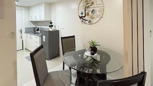 Un comedor moderno con una mesa de vidrio, dos sillas, un adorno en la pared y una cocina al fondo.